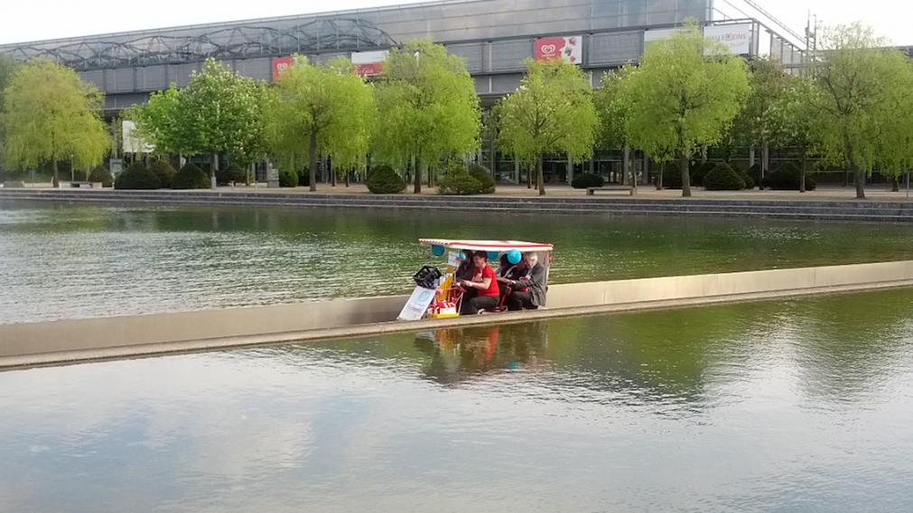 Stifter im Quandem (vierrädriges Fahrrad mit Dach) durchqueren die Wasserfläche vor der Glashalle der Leipziger Messe. Foto: Axel Halling, Berlin